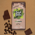 A bar of Seed and Bean Vegan Coffee 'Milk' Chocolate on a brown background with coffee beans and squares of bitten chocolate.  